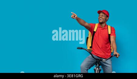 Smiling Black man pointing finger away carrying backpack sitting on bicycle against blue background in studio Stock Photo