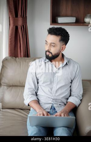 Arab man with beard working on laptop and looking sideways while sitting on couch at home Stock Photo