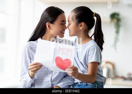 Asian woman standing and doing nose to nose with daughter holding greeting card while sitting on kitchen counter at home Stock Photo