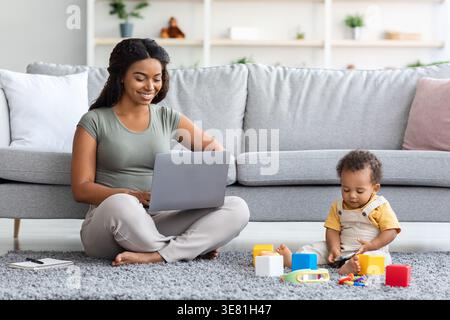 Black mother using laptop sitting with crossed legs on floor carpet near baby boy playing with his toys in living room at home Stock Photo