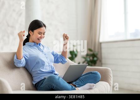 Cheerful Asian woman with clenched fist looking at laptop sitting on couch with crossed legs in living room at home Stock Photo