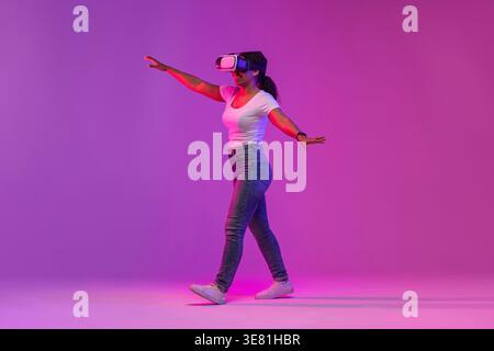 Black woman wearing white shirt and VR headset experiencing virtual reality while walking with outstretched arms against purple background with pin... Stock Photo