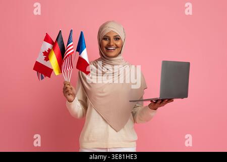 Asian woman in hijab holding laptop and international flags while standing against pink background in studio Stock Photo