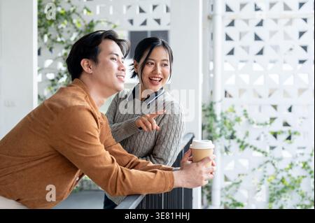 Asian man leaning arm on railing holding takeaway coffee while standing with his female friend on balcony during sunny day Stock Photo