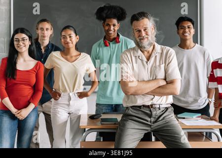Smiling diverse group of students standing behind middle-aged caucasian male teacher sitting on table in the classroom Stock Photo