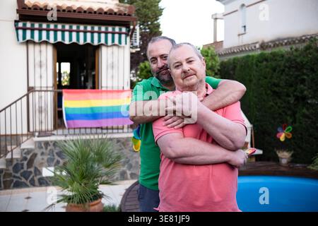 Smiling middle-aged Hispanic man embracing senior caucasian husband from behind while standing in yard near house decorated with rainbow flag durin... Stock Photo