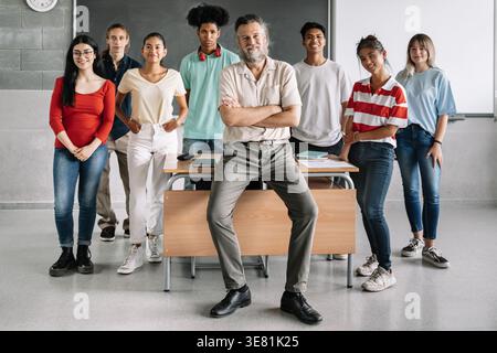 Smiling diverse group of students standing behind middle-aged caucasian male teacher sitting on table in the classroom Stock Photo