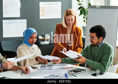Group of students taking test paper from caucasian female teacher while sitting at table in classroom Stock Photo