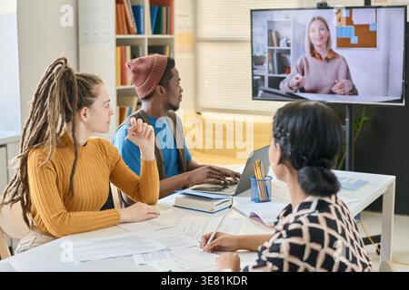 Group of students having online lesson with caucasian woman speaker while sitting at table in class Stock Photo