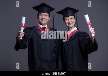 Smiling Chinese senior couple wearing black graduation gowns and caps standing with raised arms holding diplomas against gray background in studio Stock Photo