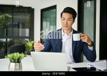 Asian businessman having online meeting on laptop holding coffee cup sitting at table in office Stock Photo