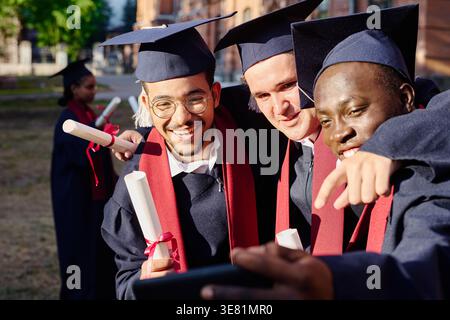 Diverse male students wearing graduation gowns and caps holding diplomas taking selfie using smartphone while standing close together on university... Stock Photo