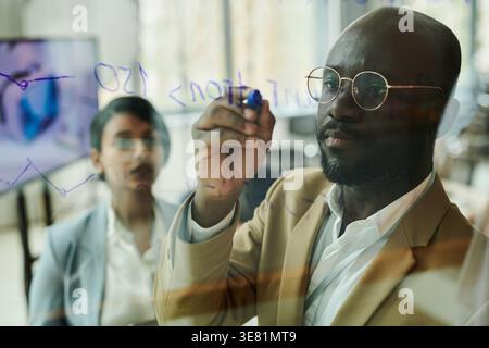 Black businessman wearing eyeglasses writing on glass in office Stock Photo