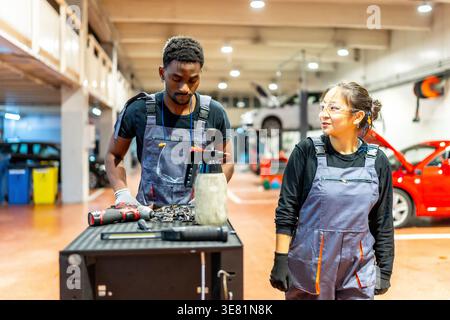 Black male and Asian female mechanics reviewing tools on workbench and discussing car maintenance during work in auto repair shop Stock Photo