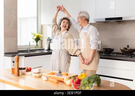 Cheerful senior Chinese couple in apron dancing near countertops in kitchen at home during daytime Stock Photo