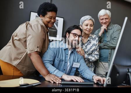 Diverse group of senior people looking at computer with caucasian male teacher while standing around him in office Stock Photo