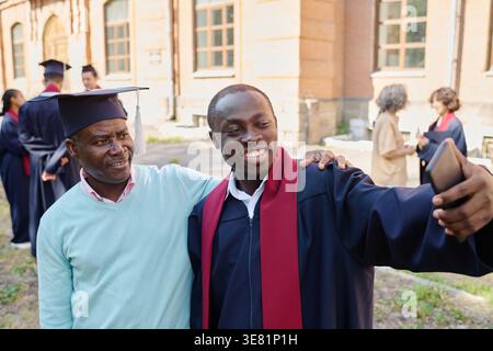 Smiling Black male student in graduation gown using smartphone taking selfie with his father wearing graduation cap while standing close together o... Stock Photo