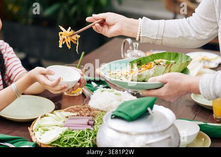 Cropped image of Asian middle-aged woman putting salad in bowl of her granddaughter while sitting at table with food and drinks in restaurant Stock Photo