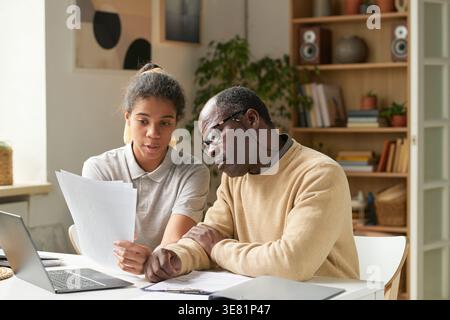 Middle-aged Black father wearing eyeglasses listening to his daughter holding documents and discussing bills while sitting together at desk at home Stock Photo