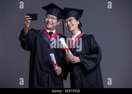 Smiling Chinese senior couple wearing black graduation gowns and caps standing holding diplomas while taking selfie against gray background in studio Stock Photo
