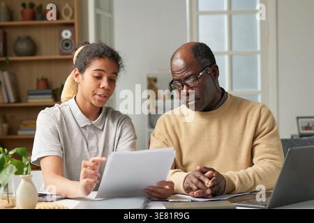 Middle-aged Black father wearing eyeglasses listening to his daughter holding documents and discussing bills while sitting together at desk at home Stock Photo