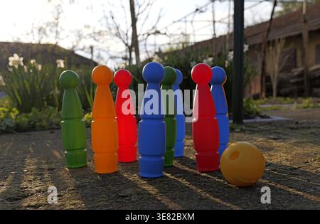 Colorful plastic bowling pins arranged in a triangle on a rough concrete surface outdoors, with strong sunlight casting long shadows in a garden. 2026 Stock Photo