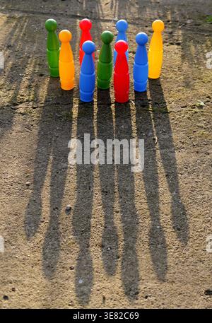 Colorful plastic skittles arranged in two rows on a concrete outdoor surface, casting long sharp shadows under strong sunlight on a clear day 2026 Stock Photo