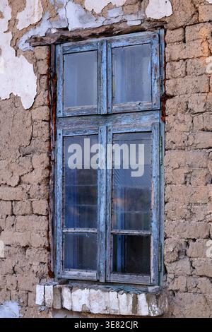Old wooden window with peeling blue paint set in a deteriorating adobe wall, showing exposed clay bricks and aged beams with strong texture details Stock Photo