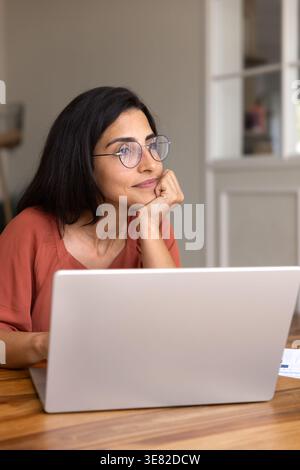 Thoughtful woman looking away seated at desk with open laptop Stock Photo