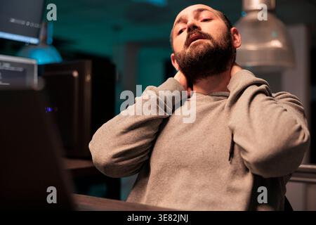 Tired male coder dealing with back pain and pressure, working late at night and feeling tense. Exhausted stressed programmer coding html script and feeling overworked, neck tension. Stock Photo