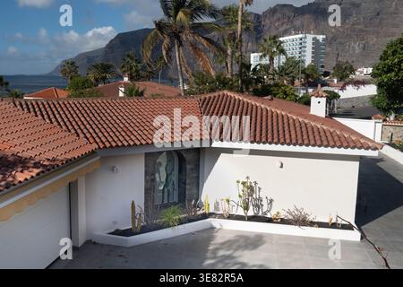 A white Canarian residential villa with traditional red terracotta tile roof, photographed from an elevated angle revealing the rooftop layout, at Los Gigantes, Tenerife, Canary Islands, Spain. The dramatic Los Gigantes sea cliffs rise prominently in the background, with palm trees and tropical garden succulents surrounding the property. Photographed in bright midday sunshine. No model or property release held â€” editorial use only. Stock Photo