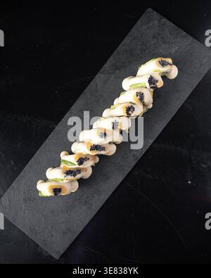 Elegant sushi roll rests on dark stone platter. Black caviar and green garnish add visual contrast. Clean lines and minimal lighting highlight gourmet Stock Photo