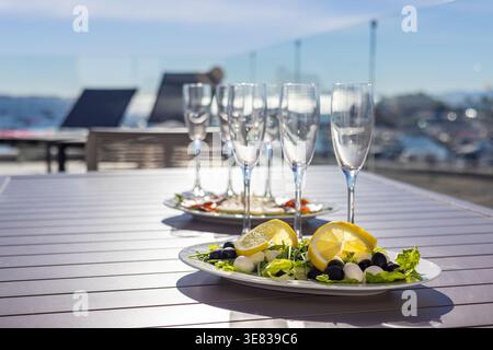 Fresh salad with olives and lemon served on a terrace table with wine glasses and city view, creating a bright outdoor dining atmosphere. Stock Photo