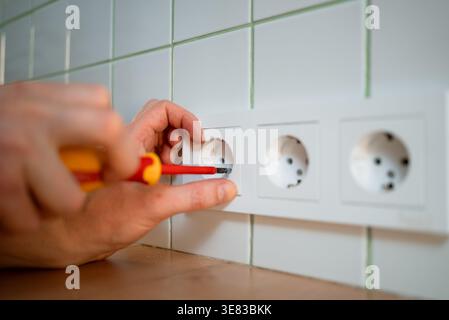 Close-up of a professional electrician's hands using a screwdriver to install a new white wall power outlet. Shallow depth of field focuses on the repair work. Home renovation maintenance concept. Stock Photo
