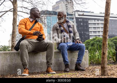 African American friends sitting and chatting on concrete bench in urban park, holding smartphones Stock Photo