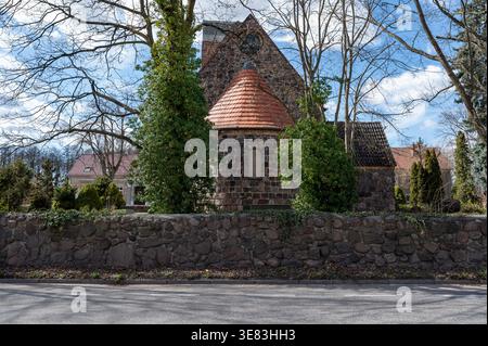 Historic fieldstone church in Schwanebeck, Barnim, Germany, with a rounded apse topped by a red‑tiled conical roof. Trees, stone wall. Stock Photo