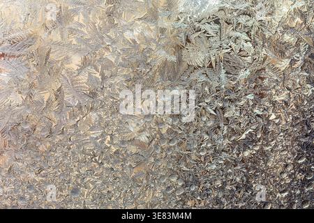 Delicate frost crystals form intricate patterns across a glass surface, shimmering in soft light. Icy texture creates a natural mosaic of silver and p Stock Photo