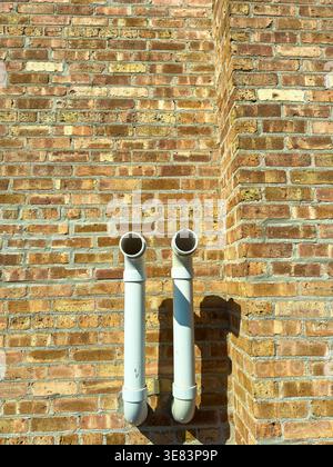 Two white pipes are mounted on a brick wall. They are positioned close together and cast shadows on the wall. The bricks are brown and warm under the Stock Photo