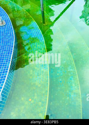 Water reflects blue tiles in a public pool. Green tones fill the water as sunlight shines. The design features steps leading into the pool. Trees are Stock Photo
