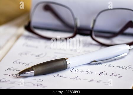 Close up of handwritten algebra equations on paper with a white ballpoint pen and blurred glasses in the background illuminated by soft natural light Stock Photo