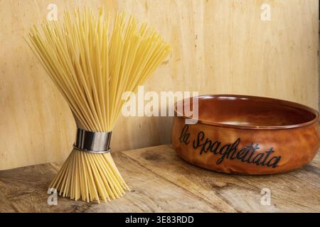 Raw spaghetti pasta tied with a stainless steel portioner on a wooden table next to a traditional Italian ceramic bowl in soft natural light following Stock Photo