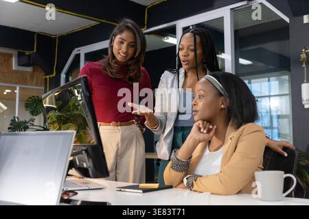 Diverse female coworkers in blazers collaborating around desktop monitor at office desk with laptop Stock Photo
