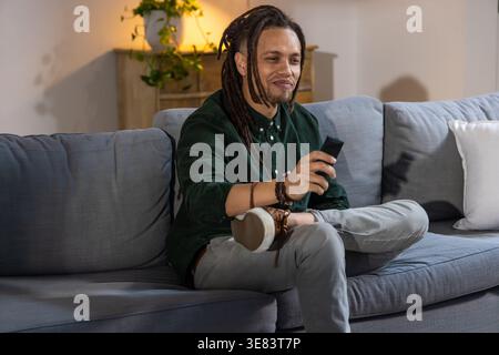 African American man sitting on sofa at home wearing dark green shirt holding black remote control Stock Photo