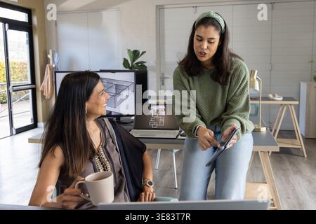 Female colleagues collaborating at desk in design studio using tablet, laptop and monitor Stock Photo