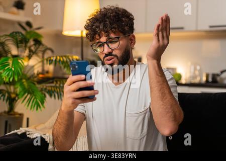 Arabian man at home checks smartphone gets bad news game loss lottery frowns shakes head no sighs. Egyptian guy on sofa reads message about result looks upset frustrated sad reacts to negative alert. Stock Photo