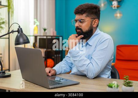 Indian man at home types on laptop stops rubs chin thinking over vital idea creative future plan. Arabian guy at table gazes screen focused eyes show deep concentration solving problem quickly today. Stock Photo
