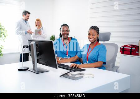 Two smiling nurses using a computer with doctors discussing in the background Stock Photo