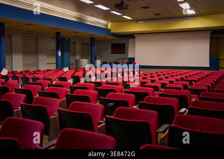 Rows of red and black chairs awaiting attendees in a large lecture hall with a blank projector screen Stock Photo