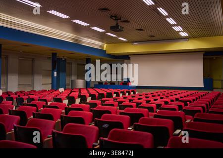 Modern auditorium hall featuring rows of empty red seats, a large projection screen, and a ceiling-mounted projector, ready for a conference or lectur Stock Photo