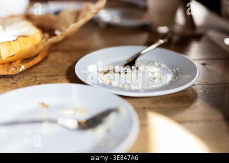 Sunlit breakfast aftermath on wooden table showing empty plates with crumbs, spoon and fork, basket with bread in soft Stock Photo